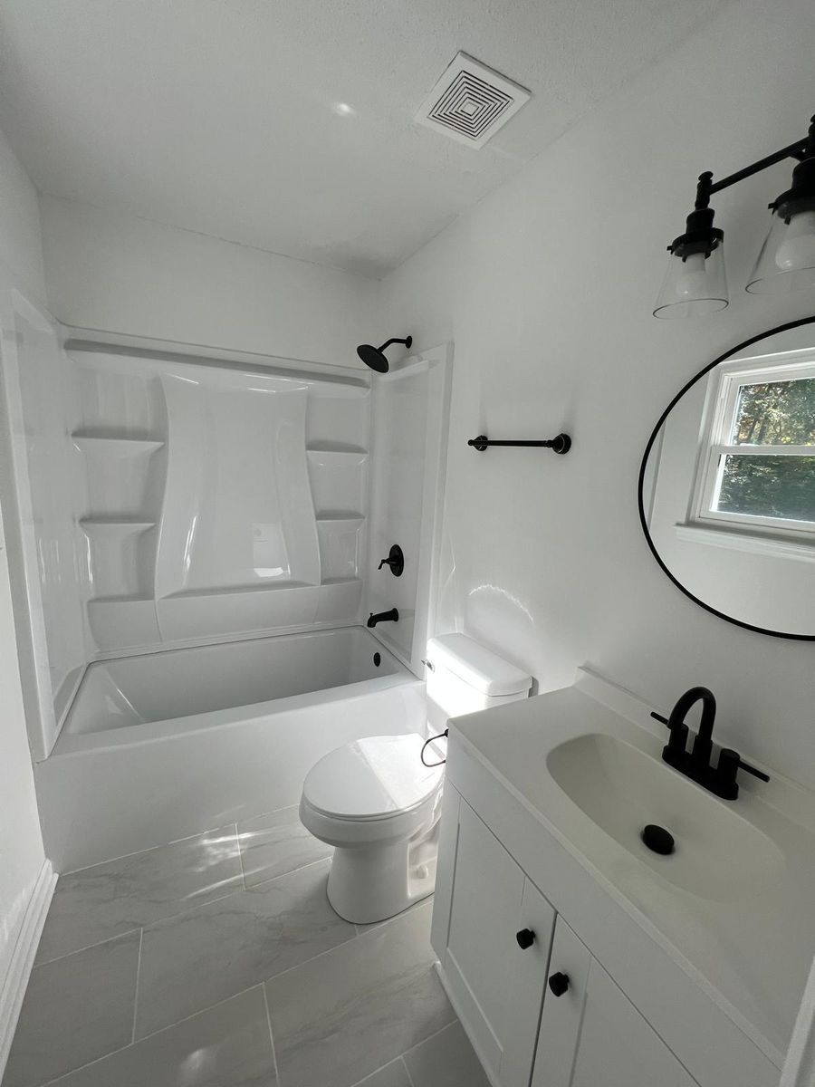 A bathroom after renovation: new tub surround, white vanity with black fixtures, round mirror, and tile floor